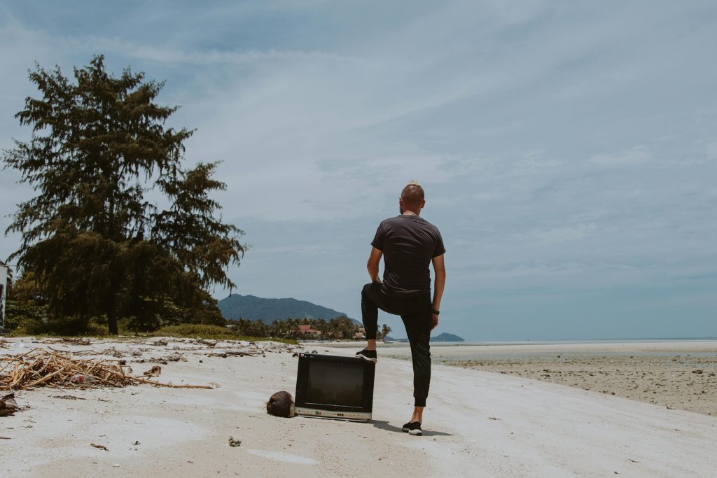 A man stands on a sandy beach next to a vintage TV, evoking nostalgia and solitude.