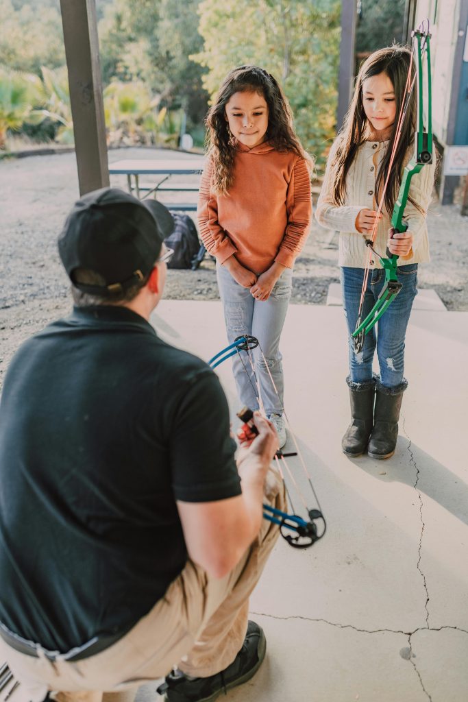 Two young girls learning archery from an instructor with a compound bow outdoors.