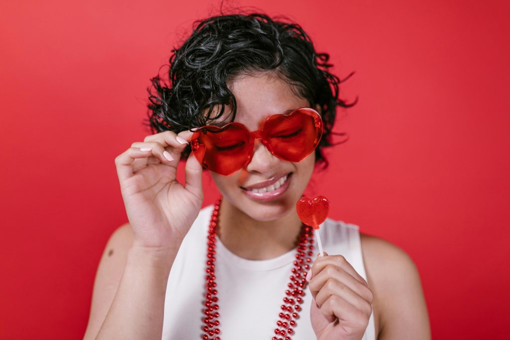 Smiling woman wearing heart sunglasses and holding red lollipop on red background.