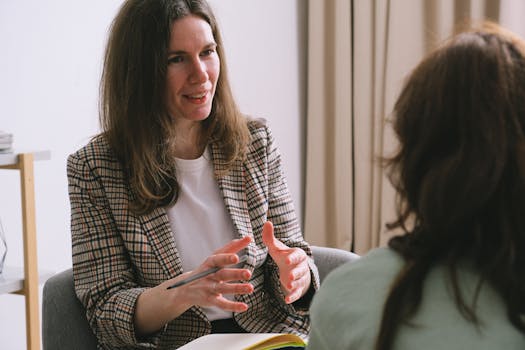 Pensive woman psychologist with brown hair in stylish clothes sitting and talking with unrecognizable female in light room